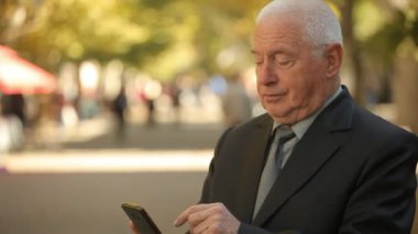 Onun smartphone bir güneşli parkta sonbahar profil net tarama ve onun Smartphone cep telefonu ile pitoresk bir parkta sonbaharda çevirme siyah bir businesssuit dayanan bir beyaz saçlı adamın içeri arama emin eski işadamı