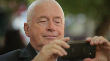 İyimser komuta sizde bir selfie onun smartphone telefon kamerasını bir selfie alarak ve sonbaharda daha iyi açı arayan uzun bir ceket bir neşeli yaşlı adamın bir yeşil sokak neşeli bokeh görünümünde taking