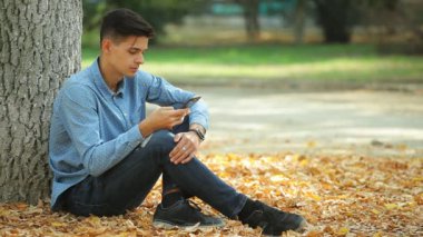 Oturma ve onun smartphone Arty profili kot pantolon ve oturma ve dalga köpüğü ağ ile bir parkta benekli gömlek yakışıklı bir genç adam, bir parkta internette tarama romantik brunet adam yaprakları sonbaharda düşmüş.