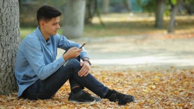 Oturma ve onun akıllı bir parkta heyecanlı profil kot pantolon ve oturma ve bir parkta telefonla düşen yapraklar sonbaharda çevirme benekli gömlek sportif bir genç adamın net sörf brunet delikanlı