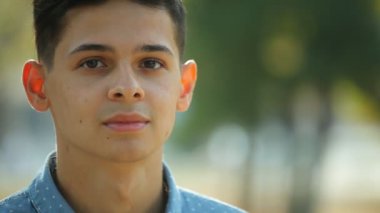 Ayakta ve gülümseyen güneşli bir sonbahar içinde neşeli bir yeşil sokak sonbahar Bokeh portre sportif genç bir gencin benekli gömlek ayakta ve güleryüzlü bir sokakta iyimser brunet delikanlı