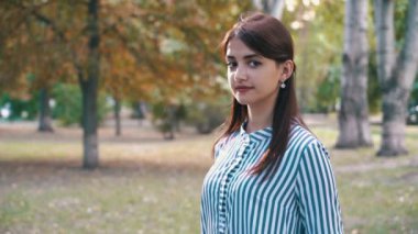Ayakta ve neşeli romantik gözleri olan genç kadının yaz üç çeyrek açık yeşil bir sokakta ve ayakta ve mutlu bir şekilde yaz aylarında yeşil bir parkta gülümseyen çizgili elbise uzun bağsız kıl gülümseyen güzel esmer kız.