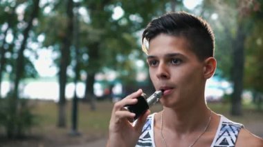 Şık brunet adam vaping yaz genç bir brunet adam portresi yeşil bir sokakta bir çizgili singlet e-sigara ve yoğun duman bir parkta streams dışarı nefes içinde mürettebat saç kesimi ile elektronik sigara