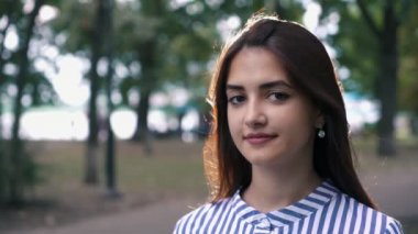 Ayakta ve romantik yaz gözleri ve uzun bağsız kıl içinde çizgili parlayan ile büyüleyici bir genç kadın portresi yeşil bir sokakta gülümseyen tatlı esmer kız elbise ayakta ve ustaca bir parkta yaz aylarında gülümseyen.