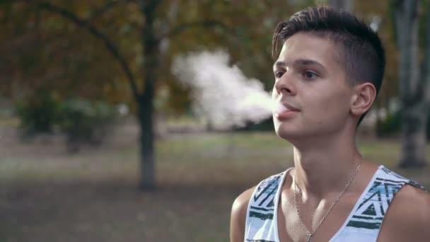 若い男が物思いにふける Vsping 電子タバコと暗い公園ストライプ一重項電子タバコを喫煙と夏の路地で密な煙を呼吸で思慮深い若い黒髪の男のプロフィールの彼の人生を楽しんで — ストック動画