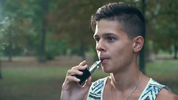 沉思的年轻人吸着电子香烟 想着他在公园里的生活 一个沉思的布鲁内特男子在条纹单打中抽着电子香烟 夏天在绿色公园里享受他的生活 — 图库视频影像