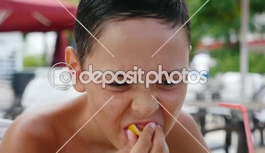 Oturma ve bir dilim limon slo-mo sıkma ve yavaş hareket yaz aylarında açık havada bir sahil üzerinde tonları ile bir kafede bir dilim limon yemek küçük bir brunet çocuk komik görünümünü bir deniz plajda bir kafede yemek küçük çocuk