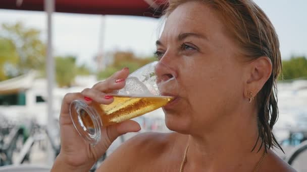一个快乐的金发碧眼的女人在海边慢动作喝着金啤酒的惊人景象 一个快乐的金发碧眼的女人微笑着 坐在户外的咖啡馆里 夏天在海边慢动作中喝着波光粼粼的啤酒 — 图库视频影像