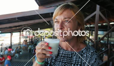 Gülümseyen kadın oturuyor ve gülümseyen ve kafeterya yavaş çekimde bir deniz plajda muzlu süt içme bir halk elbise oturuşta sahil slo-mo stylsh sarışın kadının iyimser görünümü üzerinde kafede muzlu süt içme