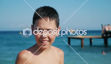 İyimser brunet çocuk vida gözleri ve kocaman bir gülümseme ile mutlu bir küçük çocuk portresi slo-mo ahşap deniz platformu gülümsüyor ve gözleri ağır çekimde yaz aylarında deniz plaj arkasında Milano platformu ile ayakta berbat etti