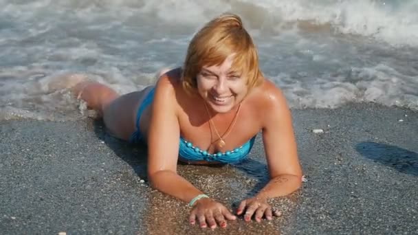 Blonďatá žena ležící na pláži moře a smilng v slo-mo v letní veselý pohled šťastné blondýnu lesklýma očima v modrých bikinách ležící na stomacn na písečné pláži v služby pěnivý vlny za slunného dne v slo-mo