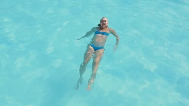 女性水泳背泳ぎおよび安静時のスローモーション水泳やスローモーションで夏は子供用プールで生活を楽しんで彼女の背中の上に横たわる青いビキニでの陽気な金髪女性の素晴らしいビューでプールをリラックス — ストック動画
