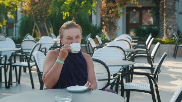 金发碧眼的女人坐在一家咖啡馆里 放松着喝着热咖啡 在懒散的时候 一个穿着黑色礼服的自信时尚的金发女郎坐在自助餐厅里 夏天喝着热咖啡 — 图库视频影像