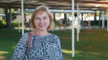 Bir teras şemsiye ile yeşil çayır üzerinde şık halk elbise ayakta bob saç kesimi ve insanlar yavaş yavaş hareket gülümseyen bir sarışın kadın neşeli görünümünü bir yeşil çim ile gülümseyen mutlu sarışın kadın