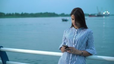 Gülümseyen ve yaz aylarında nehir iskele net tarama bir telefon ile zarif çizgili elbise uzun bağsız kıl ile çekici bir brunet kadın yaz muhteşem manzarası içinde bir nehir iskele üzerinde net sörf şık genç kadın