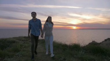 Şanslı bir adam ve onun güzel kız yaz slo-mo birlikte ağır çekimde yaz aylarında muhteşem gün batımı, Karadeniz çimenli bir seascoast üzerinde yürüme mutlu bir genç çift romantik bakış bir sahil üzerinde olacak