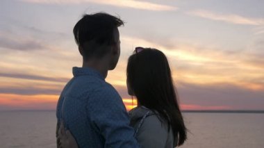 Birbirlerine kucaklayan ve ağır çekimde harika geri sportif erkek arkadaşı bir sahil üzerinde ağır çekimde yaz aylarında muhteşem gün batımı, sarılma şık bir genç kadın görünümünü batımını mutlu genç çift