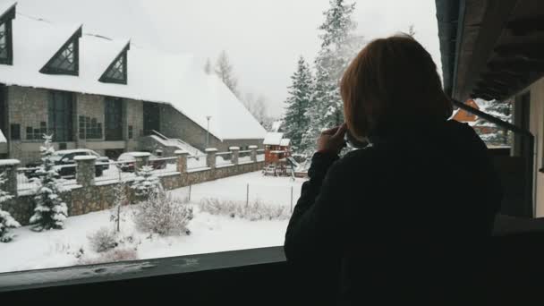 熱いお茶を飲むと山のリゾート テラスに木製の柵にもたれと雪を見てお茶を飲む毛皮のコートで思いやりのある金髪の女性のマウンテン ホテル背面ビューの雪を見て瞑想的な女性 — ストック動画