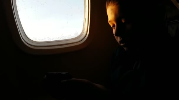 小男孩坐在飞机沙龙里 在大窗户上玩电子游戏 看到一个小男孩在智能手机上玩电子游戏的印象深刻 当时他坐在飞机沙龙里 高兴而充满了热情 — 图库视频影像