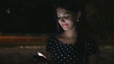Neşeli kız o smartphone ve gülümser bir sokakta net tarama ve yaz geceleri bir şehirde gülümseyen şık elbiseli uzun bağsız kıl ile yaz iyimser görünümü hoş görünümlü kız gece bakarak