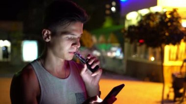 Kolsuz bluz vaping e-sigara ve kolsuz vaping elektronik sigara çabuk genç adam yaz Bokeh görünümünde net sörf ve yaz geceleri onun smartphone City hakkında bilgi arayan parlak brunet adam