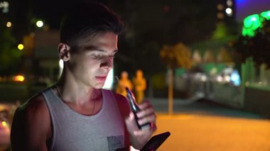 A-gömlek vaping e-sigara ve kolsuz sigara elektronik sigara çabuk genç adam yaz Bokeh görünümünde net sörf ve yaz geceleri onun smartphone şehir içinde net tarama brunet delikanlı