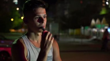 A-gömlek vaping e-sigara bir şehirde yaz Bokeh görünümünde elektronik sigara ve yaz geceleri dışarı duman City'de dudak aracılığıyla nefes kolsuz tişört rahat genç adam geceleri sokak Brunet adam