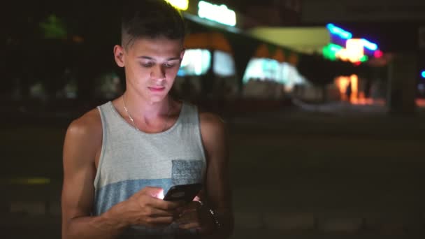 夏天的晚上 一个时髦的布鲁内男人在一个城市的街道上用手机浏览网络在他的手机上浏览夏天 在一个城市的街道上 一个穿着灰色一件衬衫的短发的黑发男人在他的智能手机上上网 — 图库视频影像
