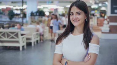 Neşeli brunet kız ayakta ve güleryüzlü bir alışveriş merkezinde yaz mutlu bir genç kadının iyimser görünümü şık beyaz elbiseli ayakta ve büyük bir alışveriş merkezinde çapraz eller ile gülümseyen uzun bağsız kıl ile çapraz kolları olan