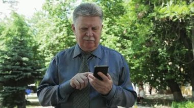 Emin beyaz saçlı adam mesaj SMS slo-mo iyimser görünümünde bir parkta gidiyor ve yavaş hareket yaz aylarında bir parkta yürürken onun smartphone üzerinde net sörf yaparken telefonunda bir mesaj yazarak akıllı ihtiyar