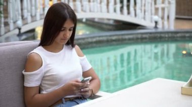 Çekici kız onun smartpnone, celeste Çeşme güzel esmer kadın şık bluz manifatura bir mesaj romantik bakış onu Smartphone cep telefonu ile muhteşem bir havuz yakınındaki bir kafeteryada bir kafede üzerinde SMS tipik