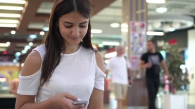 Gülümseyen esmer kadın oturuyor ve browsing belgili tanımlık enterne içinde oturan ve fotoğraflar onun Smartphone cep telefonu ile büyük bir süpermarkette kaydırma şık bir beyaz bluz gülümseyen bir esmer kızın bir alışveriş merkezi neşeli görünümünde telefonunda