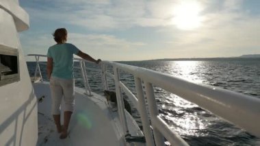 Turistik gemi yay üzerinde duran ve slo-mo Picturesque geri görünüm bir geminin yay üzerinde duran ve Mısır'daki altın güneş yolundaki slo-mo bakarak bir mavi t-shirt ve beyaz pantolon içinde bir sarışın kadın batımını ince kadın