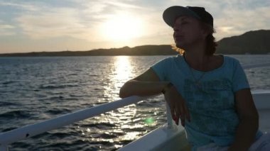 Slo-mo iyimser görünümünde bir Tshirt ve bir kap görkemli günbatımı güneş yolu slo-mo ile giymiş bir turistik gemide seyahat mutlu sarışın bir kadın ve bir gemi Küpeşte Sharm El Sheikh içinde oturan neşeli sarışın kadın 