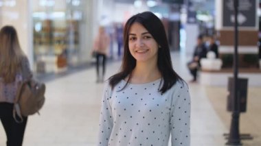 Yavaş çekimde bir bluz ayakta ve büyük pencereler ve insanlar yavaş hareket ile modern bir alışveriş merkezinde gülümseyen genç bir beyaz atlı güzellik neşeli görünümünü insanlarla geniş bir çarşıda gülümseyen çekici esmer kadın