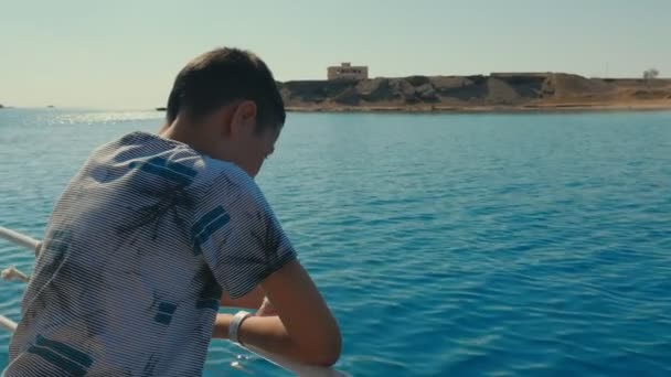 小男孩喜欢在埃及旅游船栏杆上倾斜的波涛汹涌的海浪回过头来看 一个穿着 T恤的10岁男孩靠在旅游船栏杆上 看着在阳光明媚的日子里在红海掀起波澜 — 图库视频影像