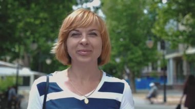 Ayakta ve gülümseyen güneşli bir günde bir gülümseyen sarışın kadın neşeli görünümünü cadde yeşille bob saç kesimi içinde duran ve bir yeşil yaz aylarında güneşli bir sokak gülümseyen çizgili bir bluz ile mutlu sarışın kadın 