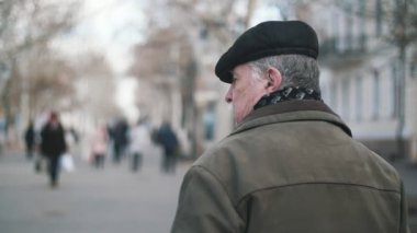 Cesur yaşlı adam beyaz bıyıklı bıyıklı düz bir kapak, yün ceket ve slo-mo kışın bir cadde boyunca yürüyüş bir Dama Tahtası eşarp slo-mo arka görünümünde üst düzey bir adam kışın bir cadde boyunca yürüyüş