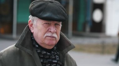 Düz bir kapak, karanlık bir eşarp ve sonbaharda bir sokakta duran sıcak bir ceket bıyıklı slo-mo neşeli görünümünde gülümseyen bir beyaz saçlı adam ve sonbaharda bir sokakta bekleyen bir kap içinde bıyıklı mutlu yaşlı adam.