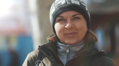 Yavaş çekimde bir örgü şapka, güzel bir ceket ve sportif bir takım elbise içinde gülümseyen bir sarışın kadın iyimser görünümü sonbaharda bir sokakta gülümseyen örme şapka neşeli sarışın kadın açık havada slo-mo sonbaharda ayakta.