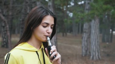 Mutlu esmer kız ayakta ve slo-mo yeşil bir ormanda e-sigara sigara yavaş hareket baharda bir çam ormanı çim elektronik sigara içen sarı kapüşonlu neşeli bir esmer kız komik görünümü