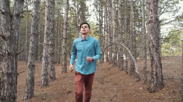 青いシャツ 茶色のパンツ 春の晴れた日に緑豊かな森の中を走っているスニーカーの陽気な若い男の印象的な眺め — ストック動画