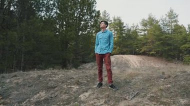 Snappy genç adam etrafa bakıyor ve slo-mo bir benekli pamuk gömlek genç bir brunet adam orijinal görünümünde baharda bir çam ahşap ayakta ayakta ayakta ve yavaş hareket baharda bir köknar ormanda bakıyor.
