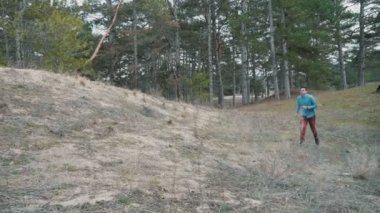 Genç sporcu esmer adam baharda çam ormanlarında ağır çekimde koşuyor pamuklu gömlekli ve kahverengi pantolonlu genç bir adamın yavaş çekimde dağlık bir çam ormanında koşuşunun ilham verici görüntüsü..       