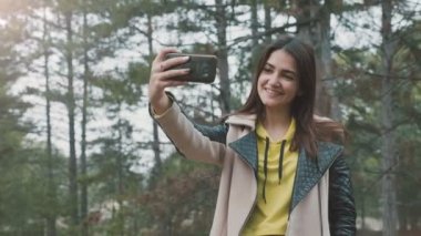 Mutlu esmer kız slo-mo uzun gevşek saçlı şık bir kızın ilkbaharda ormanda akıllı telefonuyla selfie çekerken, mutlu bir şekilde gülümseyerek, selfie çekerek, slo-mo'da ilkbaharda ormanda bir parça jest ilerler.