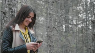 Gülümseyen esmer kız slo-mo ilkbaharda çam ahşap onu telefon net sörf genç güzel bir kadının komik görünümü onun akıllı telefon net tarama ve slo-mo ilkbaharda çam ağacında mutlu bir şekilde gülümseyerek