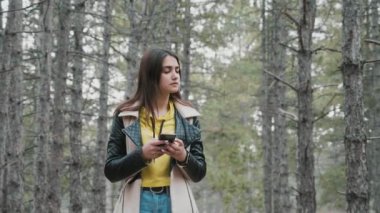 Çekici kız sağa ve sola dönerken telefonu slo-mo'da ahşap içinde. O slo-mo bir akıllı telefon ile çam ormanında kayboldu şık giysiler içinde uzun gevşek saçlı karışık genç esmer kadın heyecan verici görünümü kayboldu