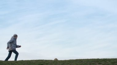Jovial çocuk çalışan ve slo-mo bir ceket, kot pantolon ve şapka neşeli bir çocuk komik profili mavi bir gökyüzü ile yeşil bir çayırda atlama, çalışan ve slo-mo bir gök gökyüzü ile yeşil bir çim mutlu bir şekilde atlama