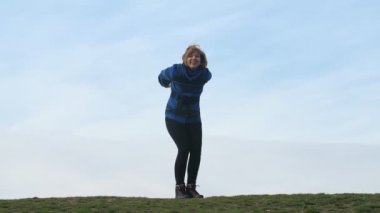 Güneşli bir günde gülümseyen sarışın kadın trendy mavi bir ceket ve kadar atlama ve bahar slo-mo celeste gökyüzü ile diz bükme tozluk slo-mo Amazing görünümünde yeşil alana atlama iyimser sarışın kadın