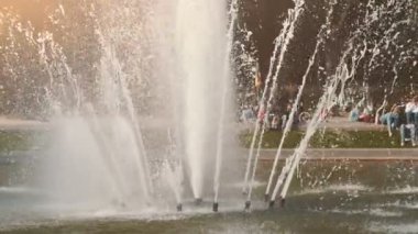 Baharda bir parkta yükselen su yüksek dereile neşeli çeşme bahar güneşli bir günde bir daire etrafında yükselen su spreyleri ile yuvarlak bir çeşme amazing görünümü. Slo-mo çim üzerinde oturan rahat insanlar
